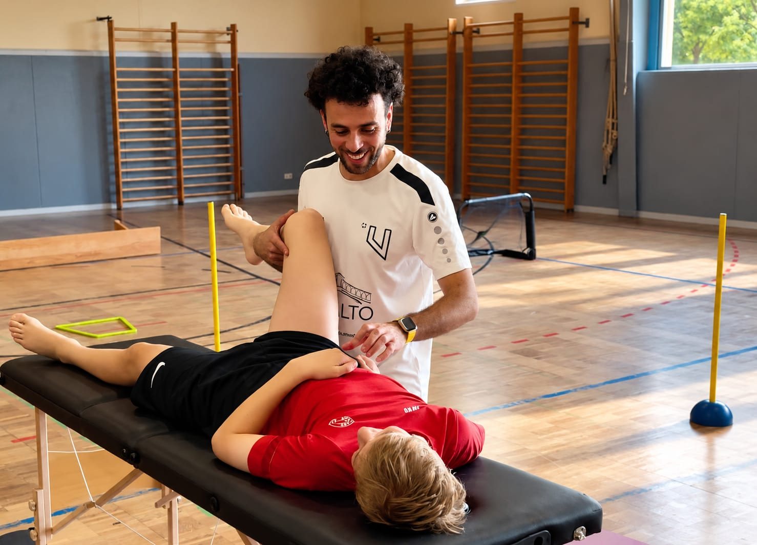 Coach Vini behandelt einen jungen Sportler auf der Therapieliege in der Sporthalle
