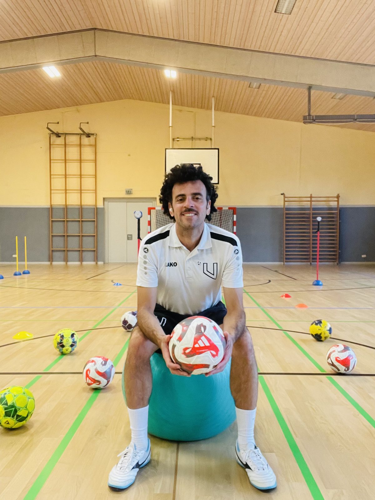 Coach Vini sitzt lächelnd zwischen Bällen und Trainingsequipment in der Halle